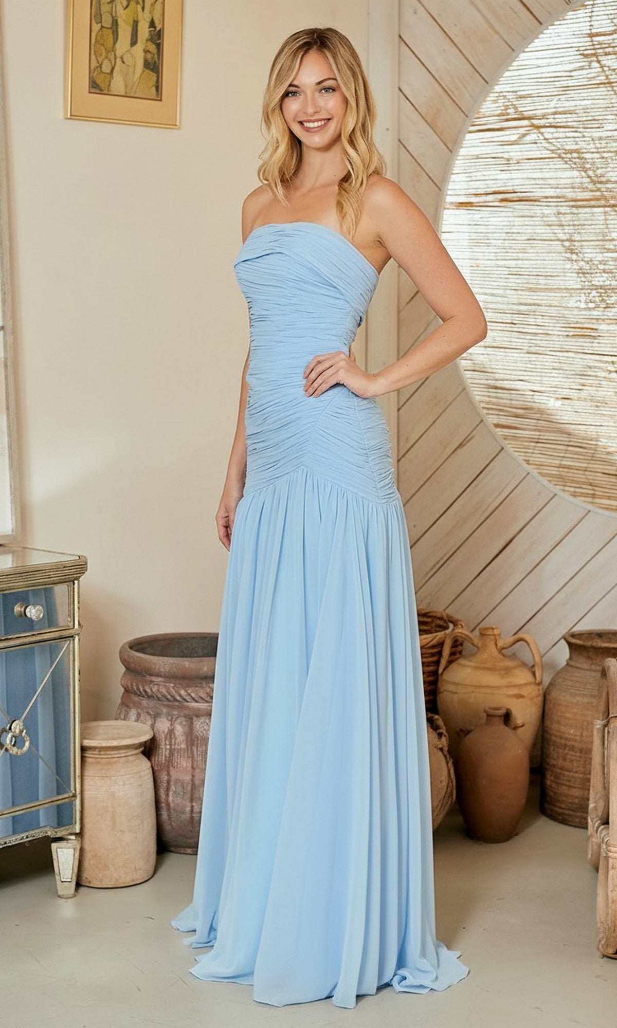 A woman in the Amelia Couture 6132 long prom dress—a strapless, light blue gown with figure-flattering ruching—smiles indoors with pottery and rustic décor in the background.